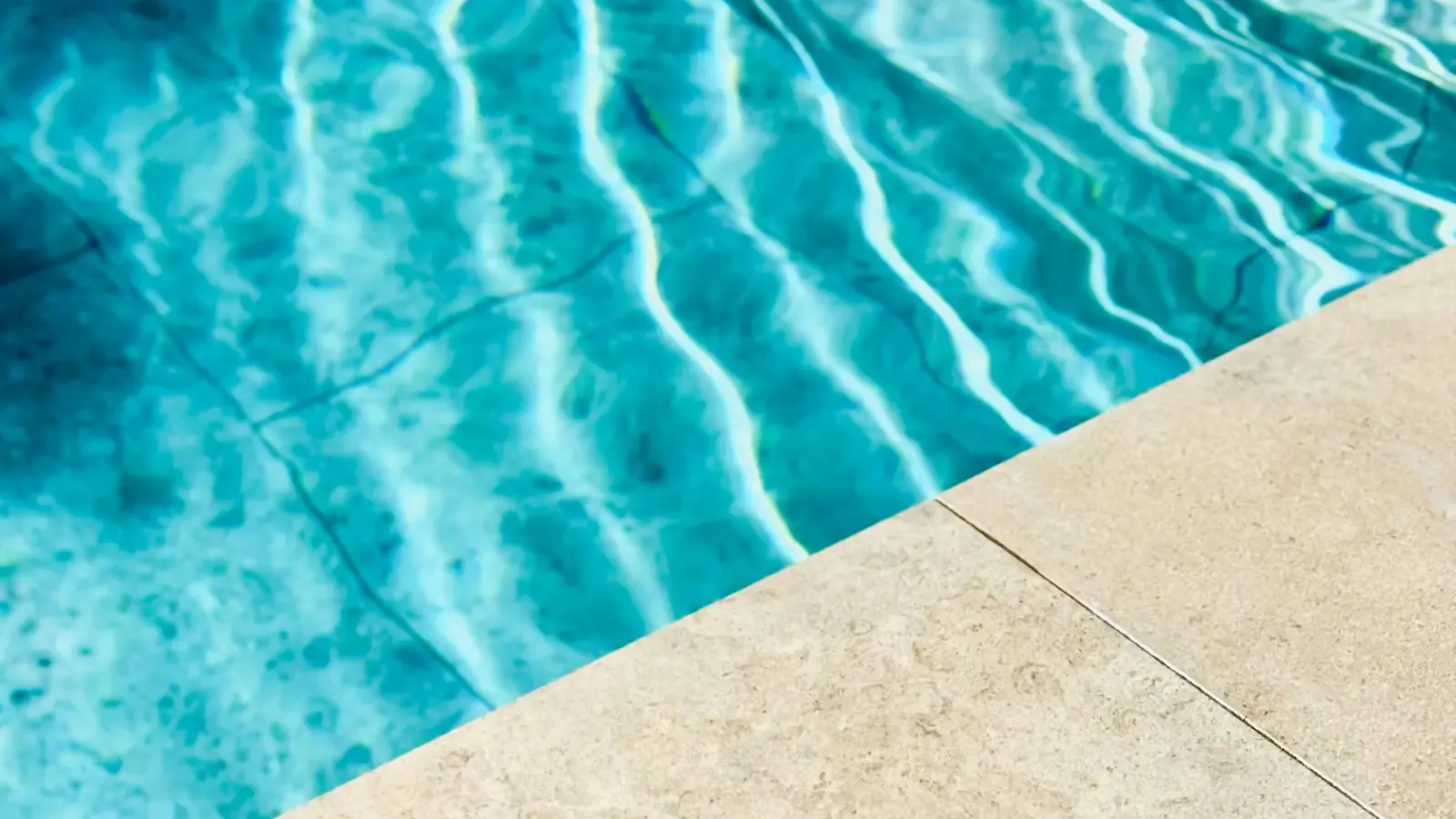 Sunlight ripples over clear turquoise pool water next to a pale stone pool edge